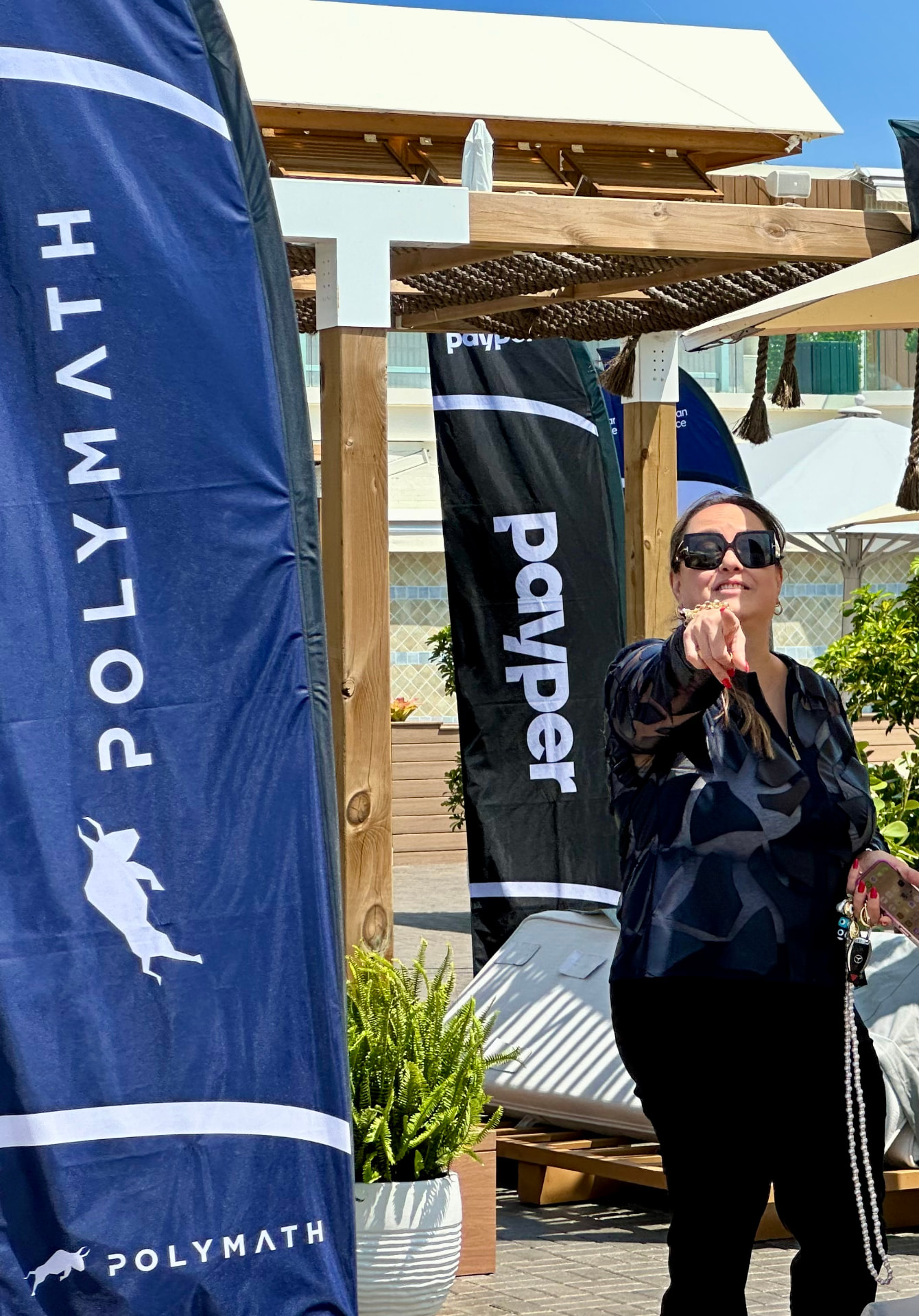 Natalie Hirsch outdoors in sunglasses beside Polymath event flags on a sunny terrace