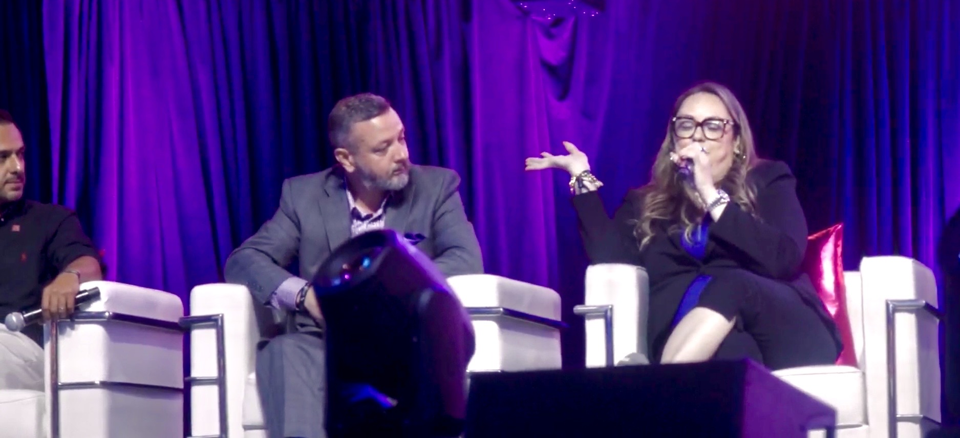 Natalie Hirsch speaking into a microphone, gesturing, on a purple-lit conference panel stage